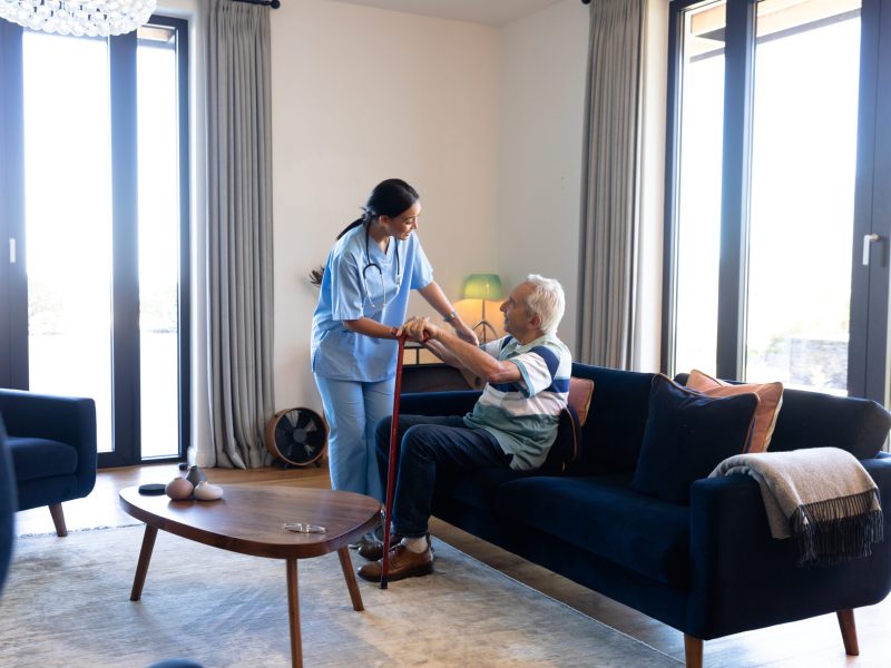 Biracial female health worker helping caucasian senior man to get up from the couch at home. Medical care and retirement senior lifestyle concept