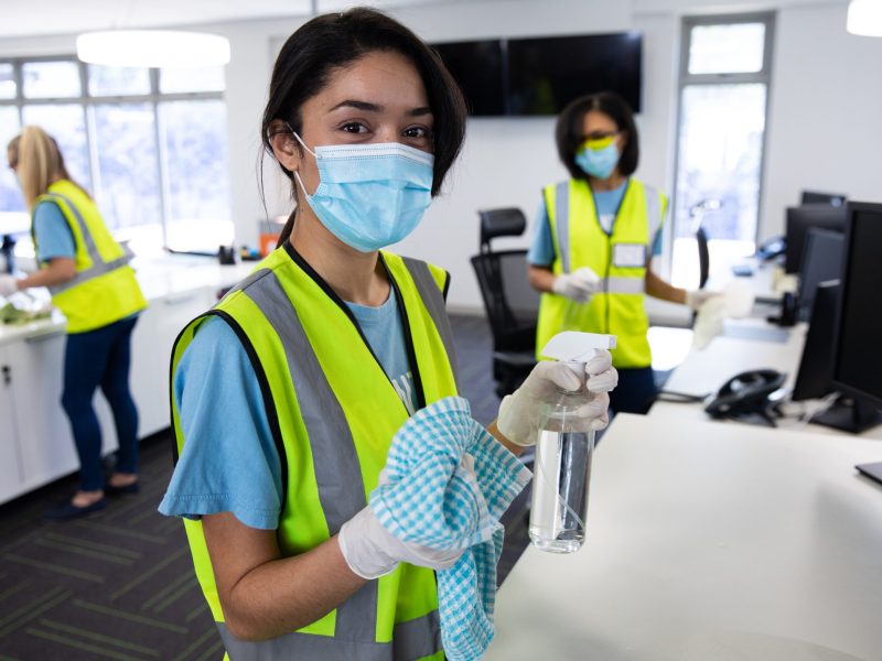 Portrait of mixed race woman wearing hi vis vest, gloves and face mask sanitizing office with disinfectant, colleagues in the background. Hygiene in workplace during Coronavirus Covid 19 pandemic.