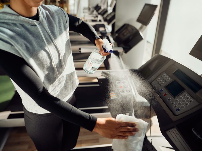 Unrecognizable athletic woman spraying treadmill with disinfectant after exercising in a gym.